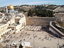 A large open area with people bounded by old stone walls. To the left is a mosque with large golden dome.