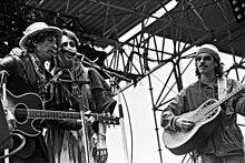 On stage outdoors, Dylan in a leather jacket and iconic porkpie hat & scarf, Baez wears a peasant skirt, sarape, tassled scarf around neck, her arm around Dylan's shoulder. Santana in workman's shirt & knit cap.