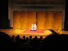 a lone person on stage sitting on a larger zabuton speaking into a microphone