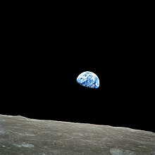 A small blue-white semicircle of Earth, almost glowing with color in the blackness of space, rising over the limb of the desolate, cratered surface of the Moon