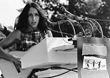 Baez stands behind a too-tall podium bristling with microphones, wearing a plaid sleeveless top, longish hair in a feather cut