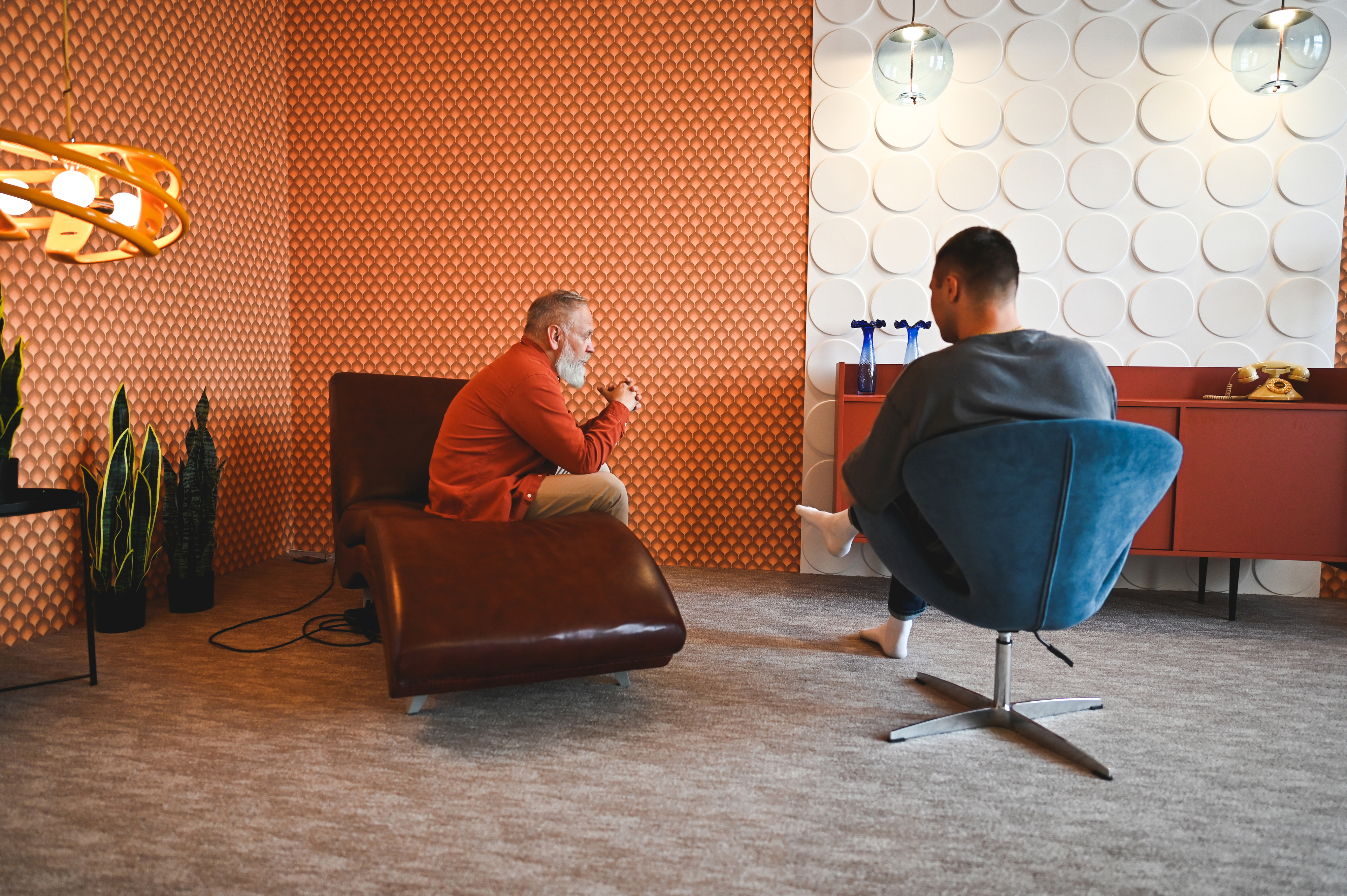 Two people sit in a stylish room, engaged in conversation. One is on a modern chair and the other on a chaise lounge, suggesting a relaxed discussion