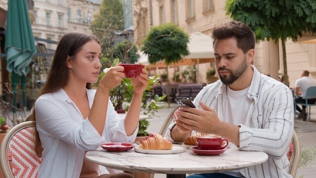 A couple sits at an outdoor café; one looks at a phone while the other sips coffee. Croissants are on the table