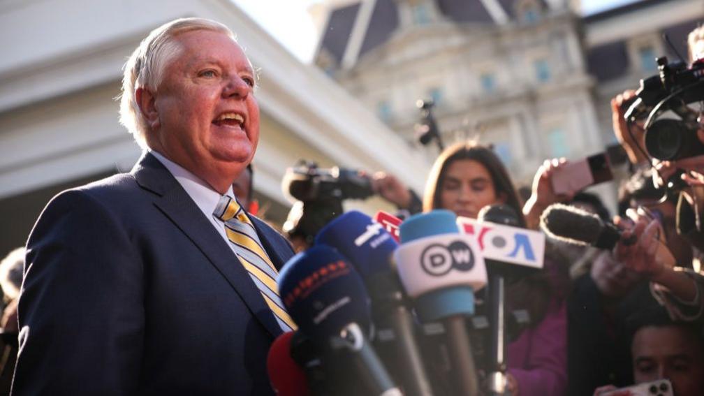 Senator Lindsay Graham in a suit and tie is surrounded by reporters outside the White House. He speaks into a microphone