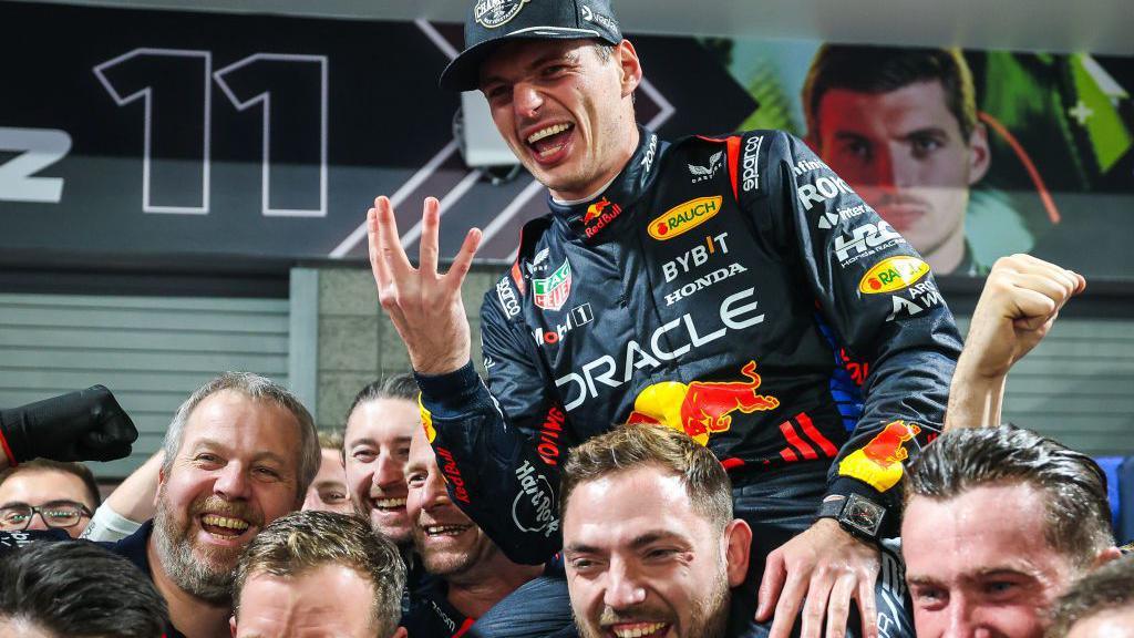 Max Verstappen holds up four fingers in celebration while sat on the shoulders of Red Bull team members