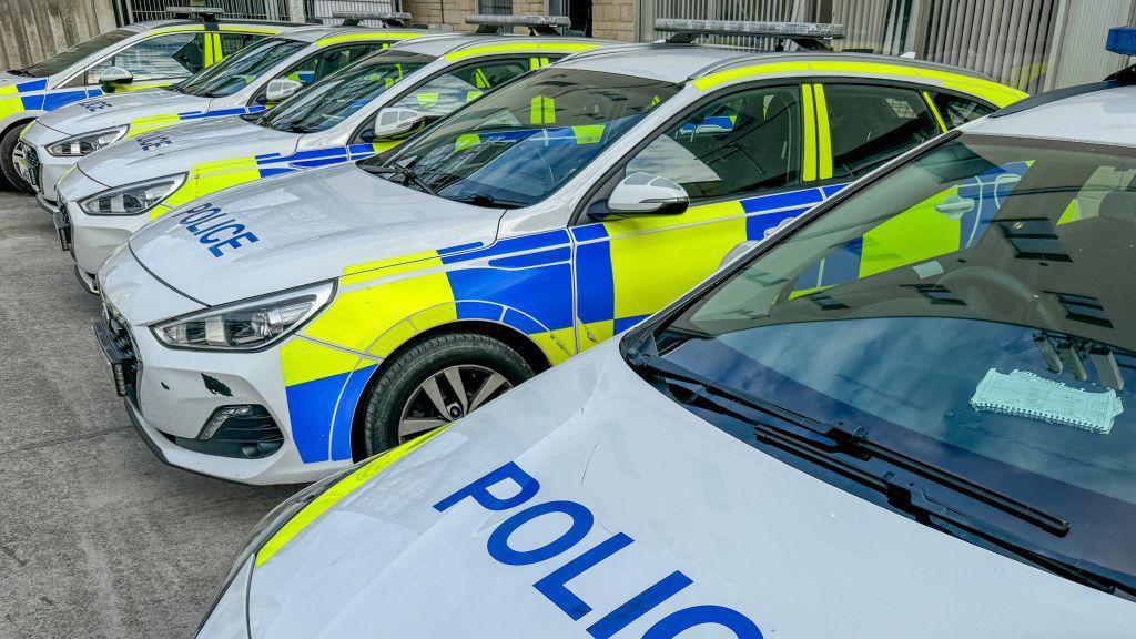 Five police cars are parked next to each other. They are white with blue and yellow patterns.