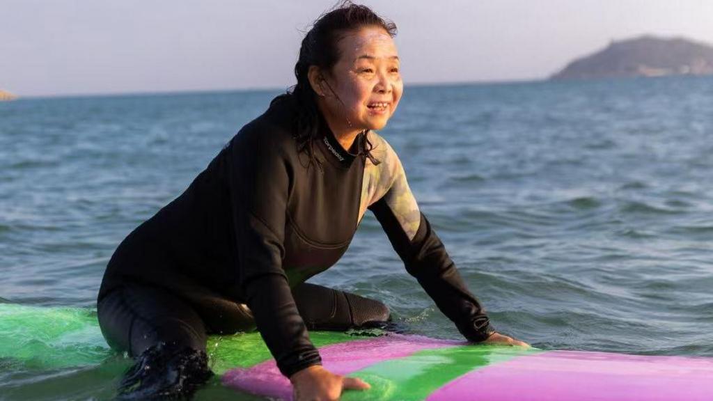 Su Min in a black wetsuit in the ocean, on a purple and green surfboard 