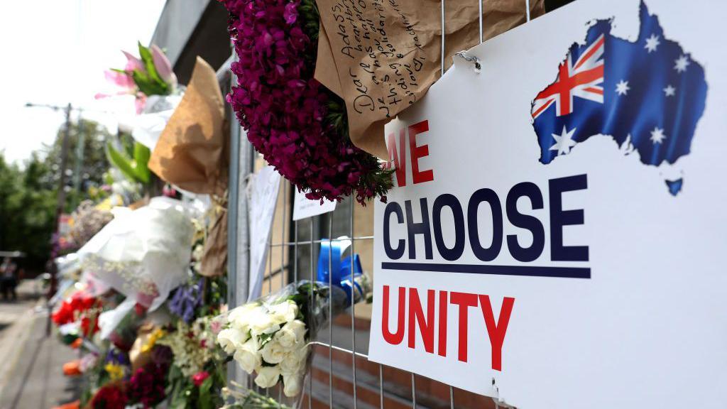 Bouquets of flowers lie in the gaps of a metal gate, which has a sign reading "we choose unity" attached