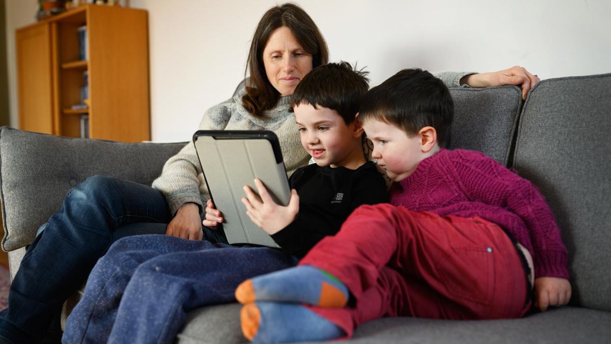 Two young children and their mother look at a tablet computer on a sofa