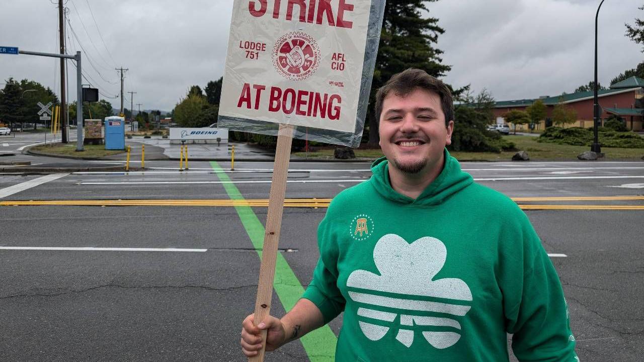 A smiling Marc Cisneros wearing a green hoody with a white clover on, standing in front of an empty road outside of Boeing holding a strike sign