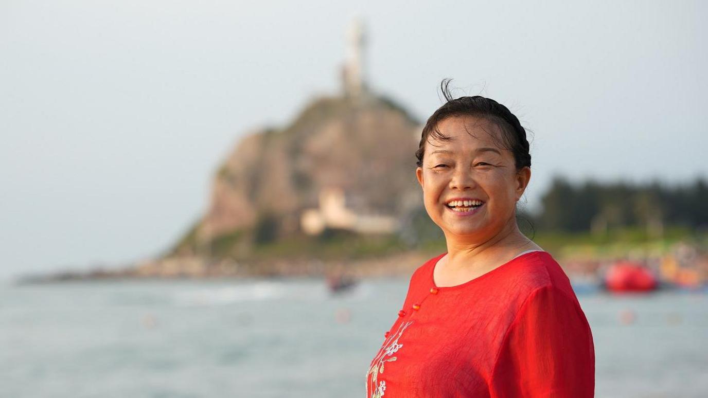 A smiling Su Min in a red top standing by a water body with a blurred hill in the background.  