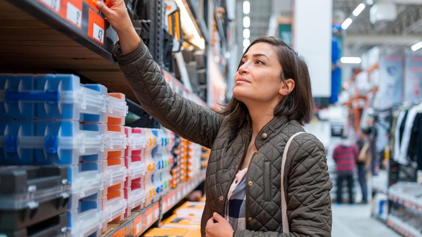 A woman with dark brown hair, wearing a green coat, shopping in a construction store. She is reaching up to a shelf which contains boxes.