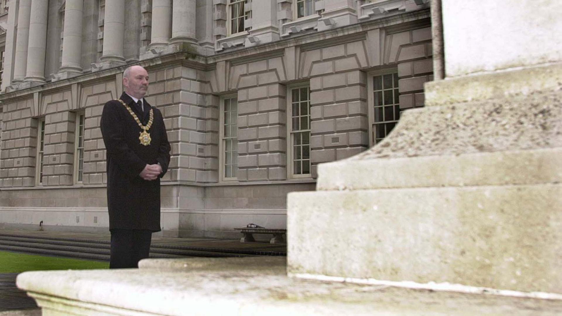 Alex Maskey stands in front of the Cenotaph outside Belfast City Hall in  July 2002.  He is wearing a long black coat, a dark suit and his gold lord mayor's chain. 