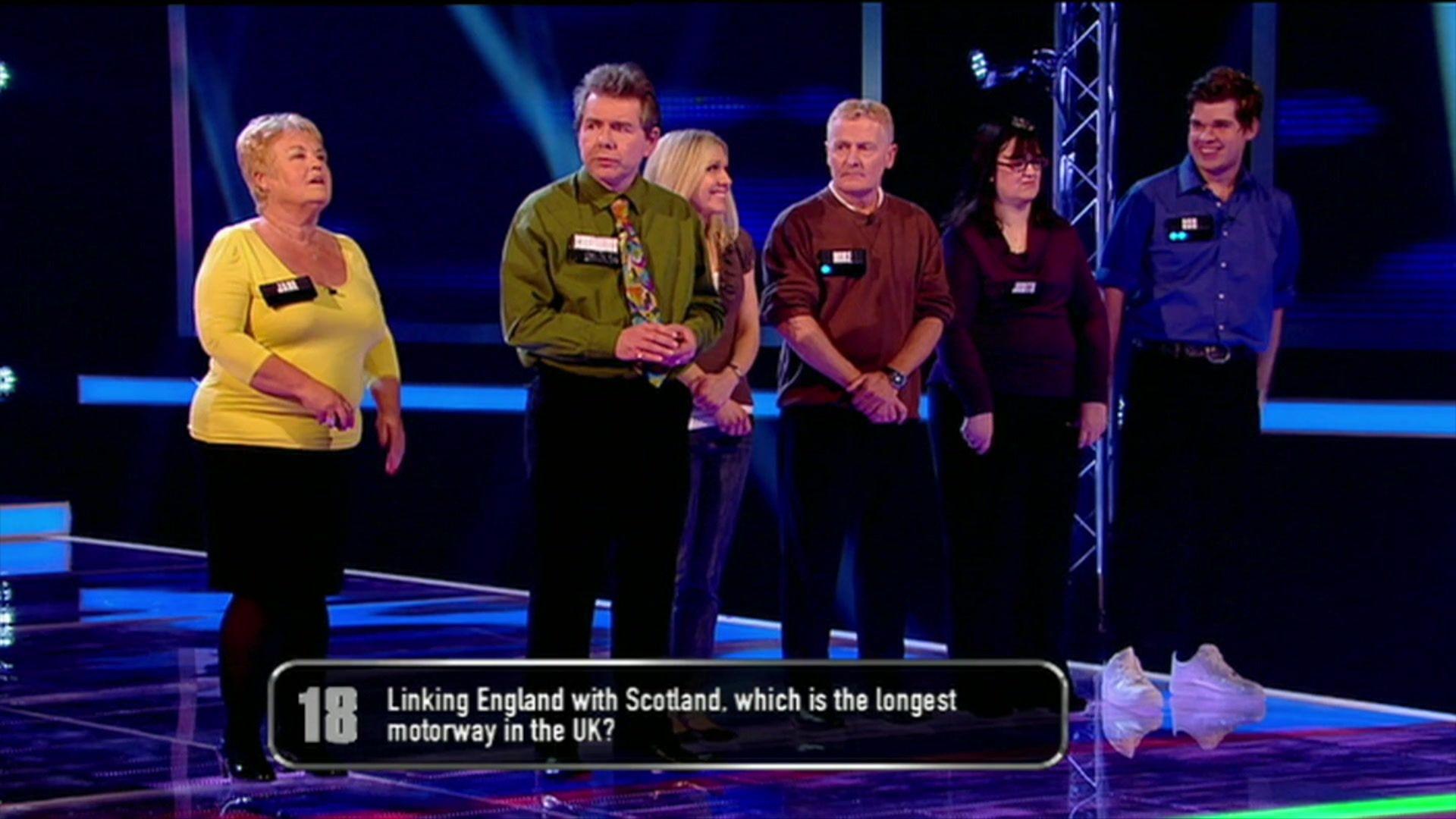 Cornelius wearing a  green shirt and brightly coloured tie standing with other contestants on Breakaway in 2012, with a question on the screen that says: Linking England with Scotland, which is the longest motorway in the UK"?