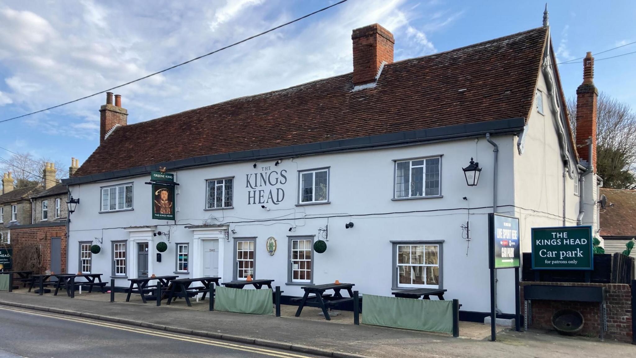 The front of the two-storey Kings Head pub. It is painted white, with brown tiles on the roof. The pub sign is of Henry VIII. Picnic tables are out the front next to the pavement.