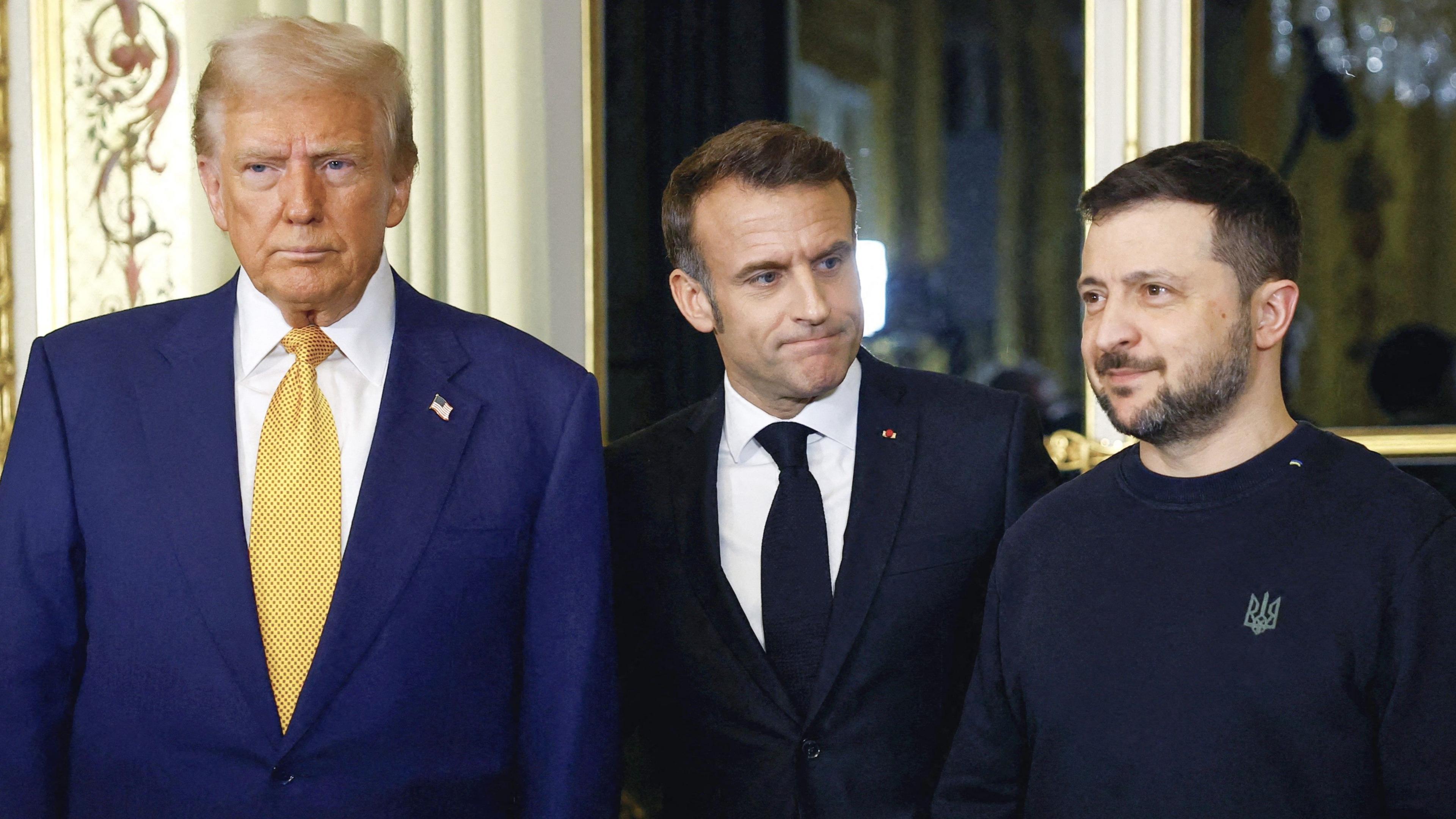 Trump, Macron and Zelensky standing next to each other during a meeting at the French Elysee Palace. Trump and Macron wear suits, while Zelensky is more casually dressed in a black sweatshirt.