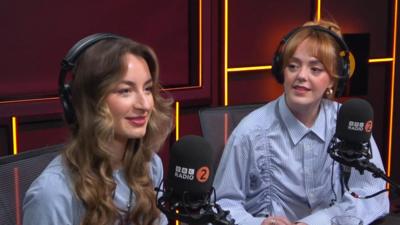 Holly and Lauren from Remember Monday sit in the BBC Radio 2 studio. They are wearing light blue shirts and tops