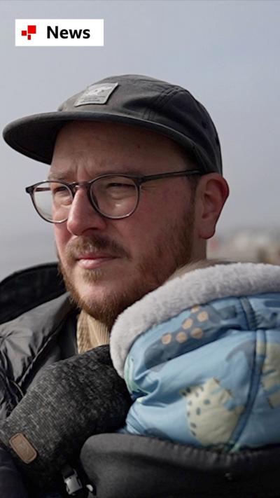 A man, Chris, holds his baby while standing at a beach. He wears a hat and glasses.