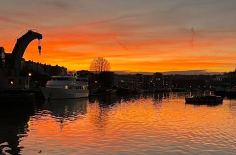 A small ferry boat travels along Bristol Harbour at sunset with an orange sky behind it and cranes and other boats silhouetted against the sky