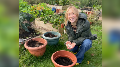 Woman is on one knee holding some white bulbs. She has blonde hair, a green coat and blue jeans. There are three tubs full of soil. In the background there are rectangular beds growing different plants. 