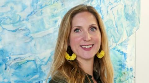 Abi Spendlove smiling at the camera in front of one of her artworks that has shades of blue representing water. She has golden long hair and wears large yellow earrings and an olive green short sleeved shirt dress.