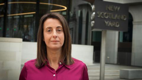 Lucy Manning, a woman in a pink blouse with brown hair, stands in front of a building looking at the camera. There is a sign that says "New Scotland Yard".