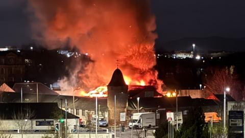 Night time shot of fire ripping through a warehouse building roof in an industrial estate. Flames and smoke are rising from the roof of the building which has a brick tower and domed roof at one corner.