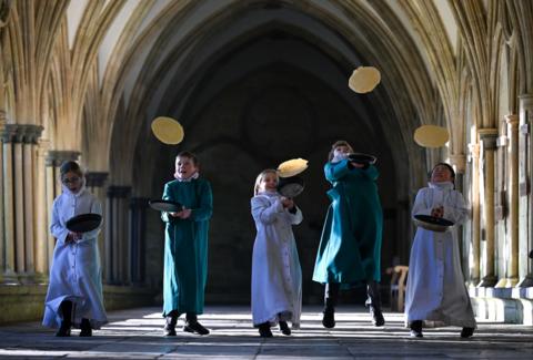 Young choristers inside a medieval cloister jumping as they flip pancakes.