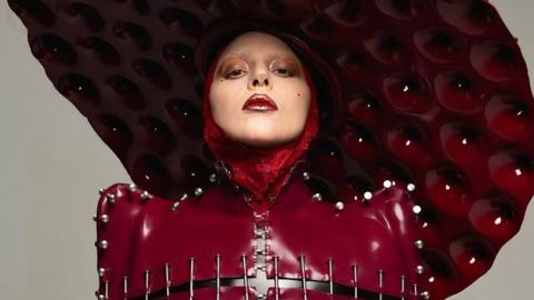 Lady Gaga wears a blood-red plastic leather outfit, stitched togeter with nails, and topped by an oversize disc-shaped hat