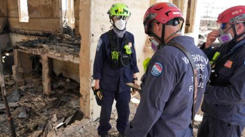 Three search and rescue team members wearing helmets and protective masks stand in a damaged building