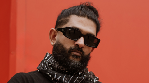 Sid Sriram, a male singer wearing black sunglasses, standing in front of a red box.