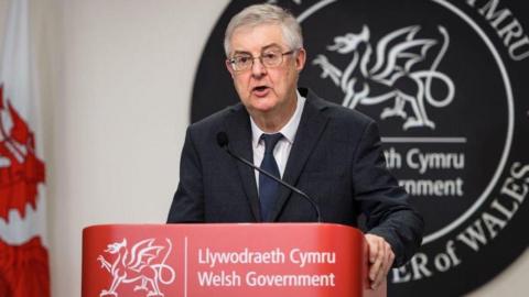 Mark Drakeford standing at a Welsh government branded lecturn hosting a news conference during the pandemic. He has a large circular grey bilingual sign made of slate behind him saying Welsh government and first minister of Wales and a Welsh flag to his right