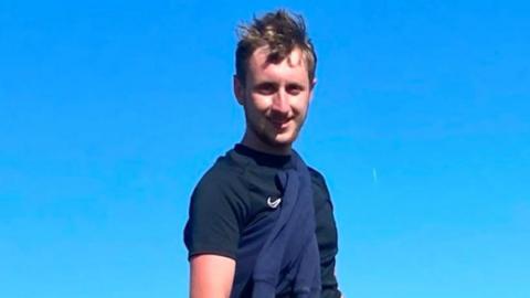 A man in a dark t shirt stands with a blue sky in the background and hills