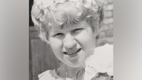 A black and white photo of a teenager or young woman. She is smiling at the camera on a sunny day and has shortish curly hair.