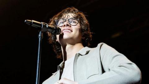 Singer David Kushner wearing black rimmed glasses and a grey shirt with a microphone in front of him, smiling and looking out into the crowd 
