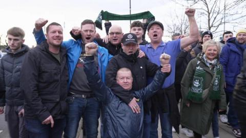 A group of Plymouth Argyle supporters sing outside the Etihad Stadium before their game away against Manchester City. A fan is holding up an Argyle scarf in the background.