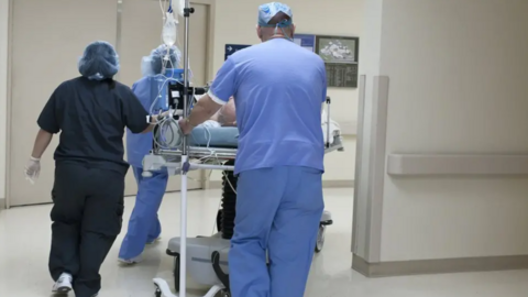 Three NHS staff members push a patient on a hospital bed along a corridor