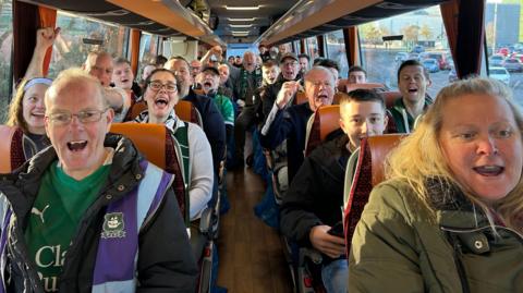 A coach full of Plymouth Argyle fans cheer loudly as they set off for a journey from Plymouth to Manchester ahead of the FA Cup tie against Manchester City. A lot of the people on the coach are wearing Argyle shirts, scarves or other clothing.