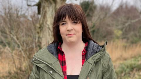 Michelle Martin wearing a green coat and plaid shirt looking at the camera with a slight smile. She is standing outside with some trees in the background.