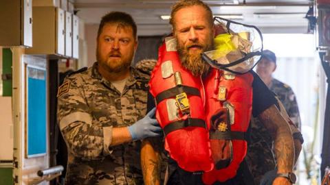 Chief Petty Officer Medic Peter Gough in military gear leads solo rowboat adventurer Aurimas Mockus, who is in an orange life jacket, to a hospital onboard the Australian warship HMAS Choules