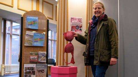 A woman dressed in a green jacket and blue jeans holds a pink bra with white polka dots above a "bra bank" - a round, pink structure with a hole in the top for depositing garments. She is standing in a library which has large windows, cream walls with gold trim and a large wooden stand holding magazines.