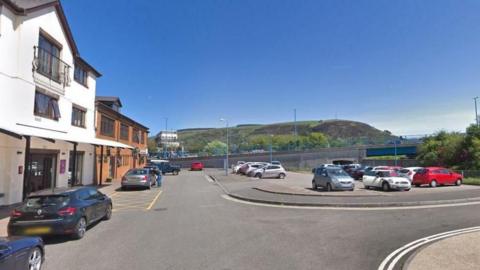 A Google street view of the car park outside Blanco's Restaurant on Green Park Street in Sandfields, Port Talbot. There are green hills in the background. A white and redbrick complex to the left. Cars are dotted all around the carpark.