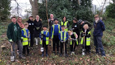 Children, parents and teachers stand in a group with shovels in wooded area