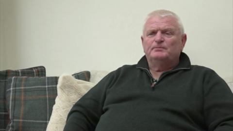 Screen grab of a man with silver hair sitting on a sofa. He looks blankly into the camera and wears a quarter zip jumper. 