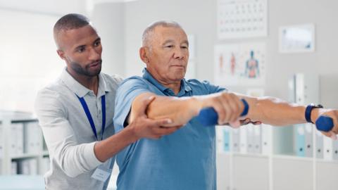 A physiotherapist standing behind an elderly man, assisting the patient with muscle training.  The physiotherapist has shaved black hair and a trimmed beard. 