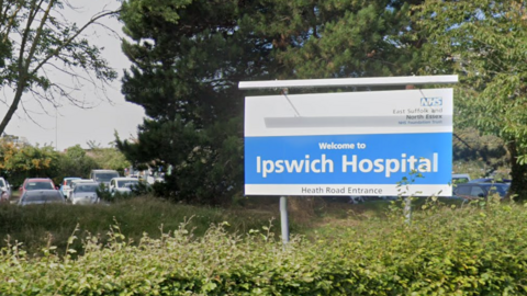 The blue and white Ipswich Hospital sign against a backdrop of greenery, including trees and bushes