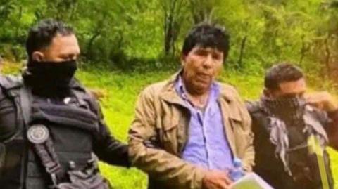 Mexican drug lord Rafael Caro Quintero is arrested in San Simon. He is wearing a blue shirt, jeans and a beige jacket. He is being held by two officers who have their faces covered.