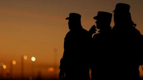 Silhouette of three Mexican soldiers at the US border. The sky in the background looks like it is sunset 