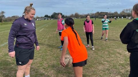 England women's rugby captain Marlie Packer talks to a girl wearing an orange t-shirt as the girl prepares to pass a rugby ball to another player. Marlie is wearing an England training jumper and shorts.