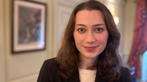 Dr Ally Louks has shoulder length brown hair, blue eyes, and is smiling at the camera. She is wearing a black blazer and white t-shirt. She is standing in a room and the lighting is casting a warm glow.